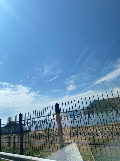 A horizontal, eye-level shot captures a serene outdoor scene with a clear blue sky dotted with wispy clouds. In the foreground, a black wrought-iron fence with decorative scrollwork runs horizontally across the frame, partially obscuring the view of the background. Beyond the fence, a calm body of water, likely Lake Baikal, stretches out, meeting a distant, tree-covered hill under the expansive sky. The grassy ground behind the fence appears dry and sparse, with faint, horizontal lines suggesting cultivated fields. A building with a reddish roof is visible to the far left, peeking over the fence. The overall atmosphere is peaceful and tranquil, characteristic of a summer day in Baikal (Port), Russia.