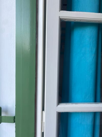 The image provides a detailed, vertical close-up of a building exterior in Φάρος, Greece. On the left, a white, textured wall transitions to a dark green, vertically-paneled wooden shutter, showing a slight dark line where it meets the window frame. A green-painted metal shutter holdback is partially visible on the lower left, attached to the white wall. Adjacent to the shutter is a white wooden window frame, appearing slightly worn with minor paint chips, particularly on its horizontal divisions. The window contains multiple glass panes, through which a vibrantly colored, solid turquoise fabric curtain is visible, drawn across the interior space. No people, actions, or interactions are present in the scene, which is illuminated by an unseen external light source.