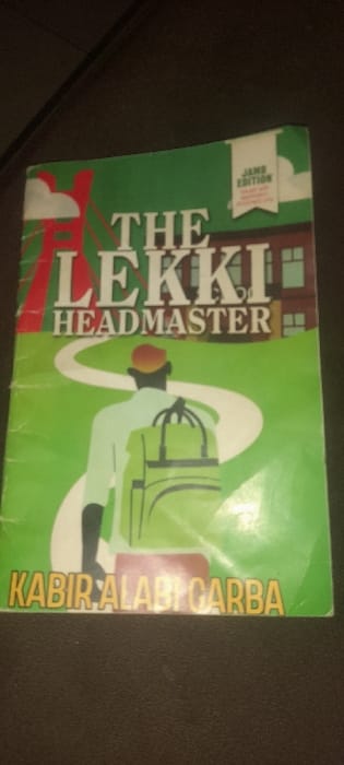 A paperback book titled "THE LEKKI HEADMASTER" by Kabir Alabi Garba is positioned face-up on a dark, textured surface in Ashipa, Nigeria. The book cover features a predominantly green background with a central white winding path. An illustrative figure, viewed from behind, wears a light-colored long-sleeved garment, dark trousers, and an orange cap, carrying a light green backpack, standing on the path. To the left of the path, a red structural element with white cloud-like shapes is depicted. To the right, a brown building with multiple windows is visible. A white banner in the upper right corner of the cover identifies it as a "JAMB EDITION." The book cover displays minor surface wrinkles and general wear. No other subjects or actions are observed in the immediate environment.