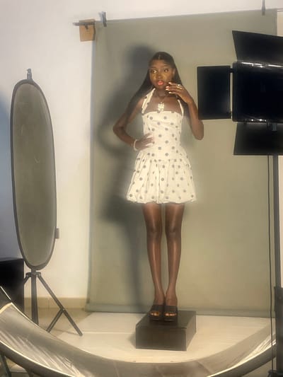A full-body shot captures a young Black woman posing for a photo shoot in what appears to be a studio. She is standing on a dark-colored cube, her body oriented vertically in the image, suggesting the photo has been rotated. She wears a short, white A-line dress adorned with navy blue polka dots. The dress features thin straps and a neckline that dips slightly. She accessorizes with a silver pendant necklace, several rings, and a delicate bracelet. Her makeup includes winged eyeliner and a vibrant pink lipstick.
The background is a plain grey studio backdrop. Above her, professional studio lighting equipment is visible, including large, rectangular lights mounted on stands, suggesting an indoor photo session. To the right, a large, translucent diffuser or softbox is positioned, further indicating a controlled lighting environment for photography. To the left and bottom of the frame, elements of studio equipment, such as light stands and a large circular reflector, are also present. The overall impression is that of a professional or semi-professional photoshoot being conducted. There is no visible text in the image. The image does not provide cues about the time of day or weather.