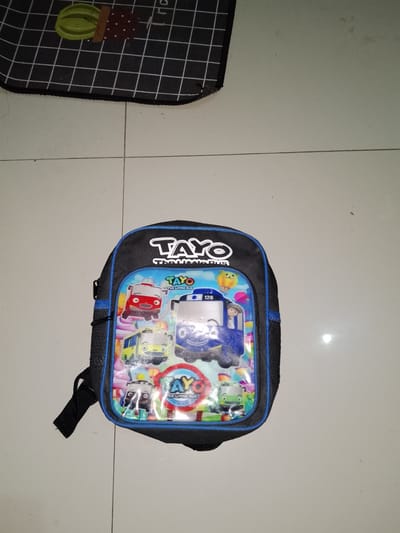 A black backpack with blue trim, featuring cartoon characters from the show "Tayo the Little Bus," lies open on a tiled floor in Sendangrejo, Indonesia. The backpack's front pocket displays images of various buses from the show, including Tayo, Gani, Lani, and Rogi, along with a round logo that reads "Tayo The Little Bus." The backpack's straps are visible, one of which is partially detached. The floor is made of light-colored square tiles, and a portion of a dark patterned mat with a cactus design is visible in the upper left corner of the image. The lighting suggests an indoor environment, possibly a room or a covered outdoor area.