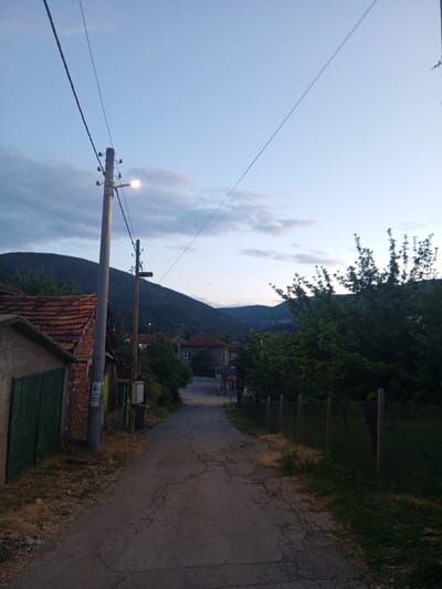 The image depicts a quiet, rural street scene at dusk in what appears to be Zemen, Bulgaria. A narrow, cracked asphalt road recedes into the distance, leading through a village or small town.
On the left side in the foreground, a building with a dark green metal gate and a visible brick wall section stands beside a tall, grey concrete utility pole. This pole supports an illuminated street light, casting a warm glow, and overhead electrical wires extend across the sky. Further down the road, more residential buildings with traditional terracotta tiled roofs are visible amidst scattered greenery. On the right, a simple wooden fence with wire mesh borders a grassy area and dense trees, which partially obscure the view of further structures.
The background is dominated by dark, undulating mountains, their silhouettes prominent against the fading light of the sky. The sky itself is a pale blue, transitioning to darker tones, with scattered clouds. The illuminated street light and the general dimness indicate that it is either dusk or early evening. A small, red hexagonal sign, likely a 'STOP' sign, is faintly visible on the left side of the road further down. No people or specific activities are visible, conveying a sense of peaceful quietude in this mountainous village setting.