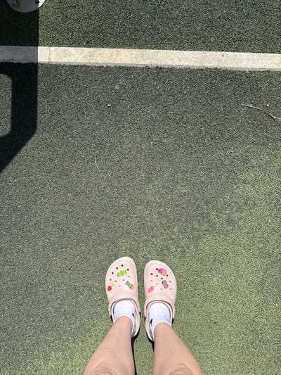 In Vyazniki, Russia, on a sunlit day, a pair of light pink Crocs, adorned with colorful Jibbitz charms resembling candy and fruit, are worn by an individual standing on a dark green, granular surface. The shoes are paired with white socks. A thin, light-colored line runs horizontally across the upper portion of the image, marking a boundary on the textured ground. A shadow cast by an unseen object falls diagonally across the upper left quadrant of the scene. The overall environment suggests an outdoor recreational area or playground.