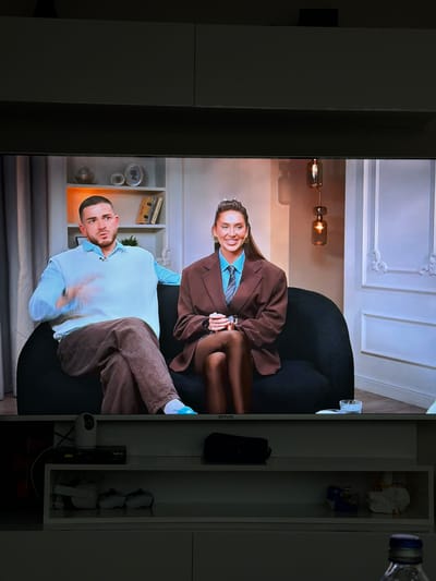 The media file shows a split screen. On the left, a person is sitting in a chair, facing the camera. They are wearing a light blue collared shirt and brown pants. On the right side of the screen, a woman in a brown blazer and blue shirt is seated, also facing the camera. Both individuals appear to be in a studio setting, possibly for a talk show or interview. The background on the left shows a bookshelf with various items, while the right background is a plain wall with decorative molding.

To the right of the screen display, there is a shelf unit with several objects, including what appears to be a bottle of water, a black bag, and what looks like gaming controllers. One of the objects on the shelf has visible text: "ONVO". Another object on the shelf has the text "DIGITURK" and "9:02".

The lighting suggests it could be daytime, or a well-lit indoor studio. No specific weather conditions are visible. The individuals' expressions are neutral, and there are no obvious indicators of a specific event or emotional state. The overall scene is calm and presented in a professional manner, likely a recorded segment for broadcast. The provided location context suggests the filming may have taken place in Kyrenia, Northern Cyprus, though this is not directly evident from the image itself.
