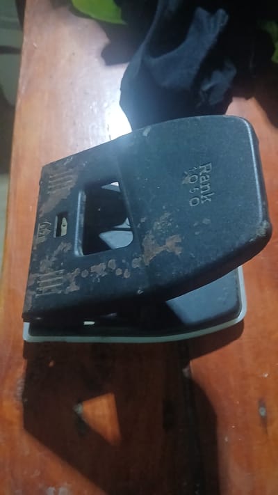A close-up shot presents a "Rank 1050" paper punch placed on a wooden surface. The punch, exhibiting signs of age and wear, with some minor corrosion marks. The scene's lighting creates a strong shadow, suggesting a single light source. Some items appear on the left of the scene, but they are heavily shadowed and cannot be described. The location of this scene is not discernible from the provided image. The details about the location in Putucual, Venezuela are not possible to determine.