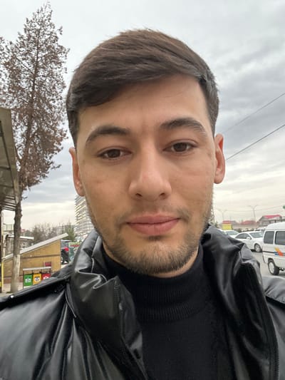 The image depicts a male individual in a close-up, outdoor setting within the city of Takhtakuprik, Uzbekistan, on an overcast day. The subject, appearing to be an adult male with light-to-medium skin tone, short dark brown hair, dark brown eyes, and stubble, occupies the majority of the foreground. He is wearing a black ribbed turtleneck sweater beneath a black, shiny puffer jacket. His expression is neutral, and he is looking directly towards the camera.
The background on the left side features a tall, leafless deciduous tree with reddish-brown branches. Below the tree, a structure with a translucent roof, possibly an awning or shelter, is partially visible. Further back and to the left, two multi-compartment recycling bins (green, red, yellow) are present near a low-rise, light-colored building with a dark roof.
The background on the right side shows an urban street or parking area. A white commercial van is prominently visible on the far right, with other white sedans parked or in motion behind it. Overhead utility lines are discernible against the light grey, cloudy sky. Distant buildings are also faintly visible on both sides of the background.