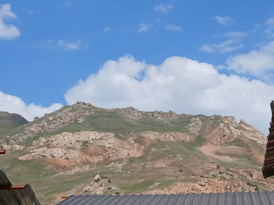 The image depicts a mountainous landscape under a blue sky with scattered white clouds. The foreground features the corrugated metal roofs of two structures, one on the left and one on the right, partially obscuring the view of the mountains. The mountains themselves are characterized by rugged, rocky slopes covered in patches of green vegetation. The terrain is uneven, with exposed rock formations and earthy tones of brown and tan interspersed with the green. The scene appears to be an outdoor environment, likely in a rural or natural setting. There are no people or animals visible in the image. The location in the city of Kanchoch, Tajikistan is noted, suggesting this is a view of the natural surroundings in that region.