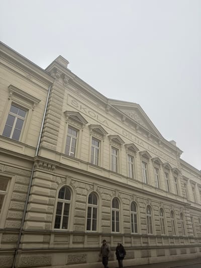 The image displays the ornate, multi-story facade of Gymnázium Dr. Karla Polesného, located at Komenského náměstí 3 in Znojmo, Czechia, under an overcast sky. The building is painted a light cream or beige. The ground floor features rusticated stonework and several arched, white-framed windows. The upper floor exhibits a smoother surface with rectangular, white-framed windows, each topped with a decorative pediment. Above the upper windows, a large inscription reads "ŠKOLA - DÍLNA LIDSKOSTI" (SCHOOL - WORKSHOP OF HUMANITY), centered below a triangular pediment with intricate relief carvings. Decorative architectural elements, including pilasters, cornices, and carved faces, are visible along the roofline. A silver-colored downspout runs vertically along the leftmost visible section of the building. Two individuals are walking from left to right on the wet sidewalk in front of the structure. The person closer to the camera wears a dark jacket and lighter trousers, while the second person, slightly behind, wears a dark coat and carries a shoulder bag. The overall environment suggests a cool, potentially damp day with soft, diffused light.