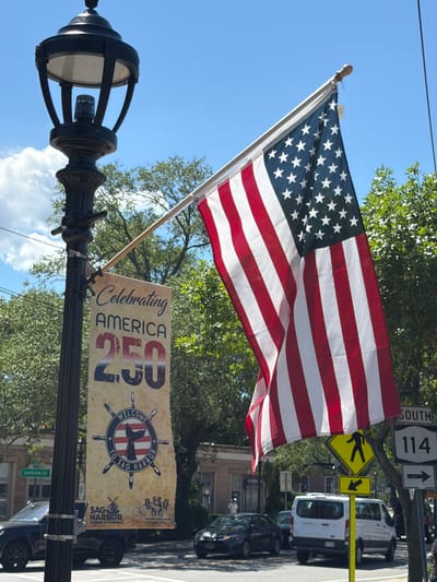 A black lamppost stands on the left side of the image, topped with a decorative lantern. An American flag, with red and white stripes and a blue union with white stars, is unfurled and attached to the lamppost via a metal bracket and pole. Below the flag, a beige banner hangs from the lamppost, displaying text and graphics. The top of the banner reads "Celebrating AMERICA" in stylized lettering, followed by "250" in large, bold numbers. Below this, a circular emblem features a ship's wheel with a whale's tail in the center, inside a circle with the text "WELCOME TO SAG HARBOR". At the bottom of the banner, logos for "SAG HARBOR CHAMBER OF COMMERCE" and "HAMPTONS GYM" are visible. The background consists of green trees, blue sky with scattered clouds, and buildings. In the distance, vehicles are on a street, including a black SUV on the left, a white van in the center, and a white car on the right. Traffic signs are also present, including a "DIVISION ST" sign and a "SOUTH 114" sign. The overall scene depicts a street in the Village of Sag Harbor, United States of America, celebrating an event.