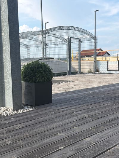 This media file captures an outdoor scene, likely during the daytime under a partly cloudy sky, suggesting fair weather. The perspective is from a slightly elevated position, looking across a wooden deck towards a large, semi-circular metal structure with a wire mesh. This structure appears to be a cage or enclosure, possibly for an animal or an event.

In the foreground, to the right of the frame, is a dark grey, cubic planter containing a neatly trimmed, spherical green bush. The planter rests on a bed of small, light-colored pebbles that extend towards the wooden planks of the deck. The deck itself is made of weathered, grey wooden planks laid parallel to each other.

To the left of the planter, behind the metal structure, elements of a building are visible. These include red-tiled roofs and part of a building with a yellow facade, suggesting an urban or suburban setting. The metal structure is supported by poles and has a complex framework.

A notable detail is a long, thin metallic arm extending horizontally from a pole on the left side of the metal structure, pointing towards the sky.

The scene is devoid of people, focusing on the architectural elements and the planted greenery. The overall impression is one of modern landscaping combined with functional or industrial structures. The location context is Singen (Hohentwiel), Germany, though no specific landmarks are immediately identifiable from the image alone. There is no visible text in the image.