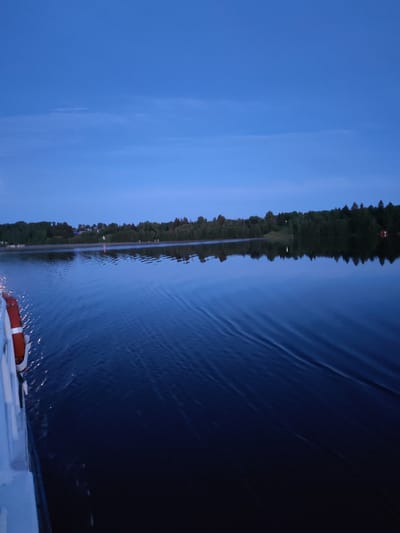 The image captures a serene twilight view of a calm lake with a distant treelined shore. The sky is a deep, clear blue, transitioning to a darker hue near the horizon. On the left side of the image, the stern of a white vessel is visible, with an orange life preserver attached to its railing. The vessel is creating gentle ripples in the dark blue water, which reflects the twilight sky and the silhouettes of trees. The shore is densely wooded, with a few scattered buildings and lights visible amongst the trees. The overall atmosphere is tranquil and peaceful, suggesting a quiet evening on the water. The location is specified as Vskhody, Russia, and the scene suggests a natural environment, likely a lake or a wide river within or near the city.