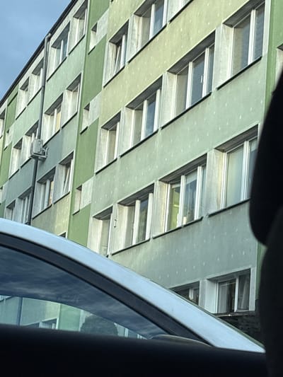A side view of a pale green, multi-story building is visible, with a white car in the foreground. The building has many windows with white frames, arranged in horizontal rows. The windows are of varying sizes and some appear to have blinds or curtains inside. A gray pipe runs vertically along the side of the building, with a small gray electrical box attached to it. The sky above is a light blue. The image appears to have been taken from inside another vehicle, as suggested by the reflection in the car's window. This scene is likely located in the city of Kalisz, Poland, based on common architectural styles and urban environments found in the region.