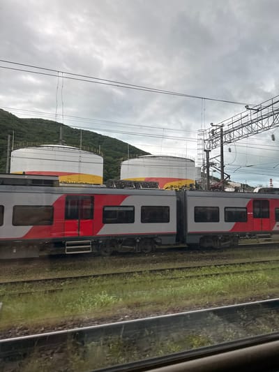 The image shows a view from inside a moving train. The train car is red, white, and gray with rectangular windows. Outside the train, there are parallel train tracks with green vegetation growing between them. Beyond the tracks are large white cylindrical tanks with yellow and red bands around the top. Behind the tanks, there is a forested hill. Overhead are electrical power lines and a gray, cloudy sky. The location is not specified within the image itself.