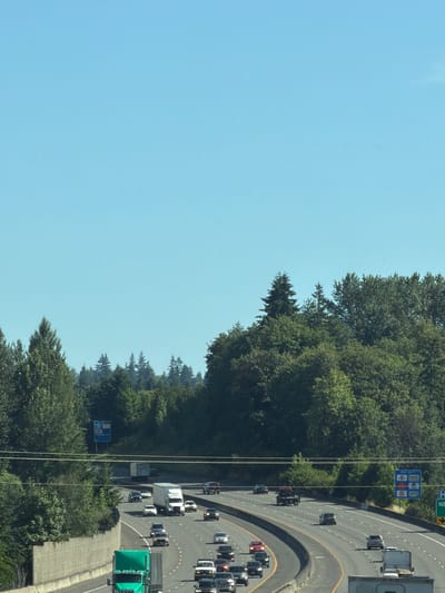 This media file shows a view from above of a highway in Vancouver, United States. The highway is curving to the right and is filled with various vehicles, including cars, trucks, and what appear to be SUVs. The traffic seems to be moving at a moderate pace. On the left side of the highway, there is a dense line of trees, forming a natural barrier. The sky is clear and blue, suggesting a sunny day. There are power lines visible in the foreground, running vertically through the image. On the side of the highway, there are a couple of blue signs. One sign appears to be a directory for services, with multiple squares depicting logos for fast-food restaurants like McDonald's and possibly other amenities. Another sign is a green directional sign with white lettering indicating "NE". The overall scene depicts a typical day on a busy highway surrounded by nature. The time of day appears to be daytime, given the bright sunlight and clear sky. There are no discernible emotions conveyed in the image, as it focuses on the landscape and traffic.