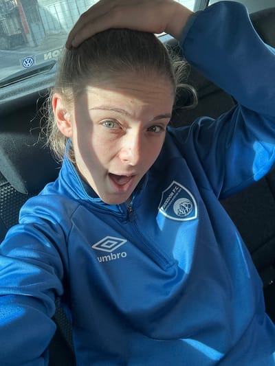 A person with fair skin and light hair, wearing a blue athletic jacket, poses in what appears to be a car. The jacket has logos for "FREEDOM F.C." and "Umbro". The person's mouth is open in a surprised or excited expression, and one hand is on their head. Part of the vehicle's window and door frame is visible, along with the shadow of the person. The overall lighting suggests a sunny day.