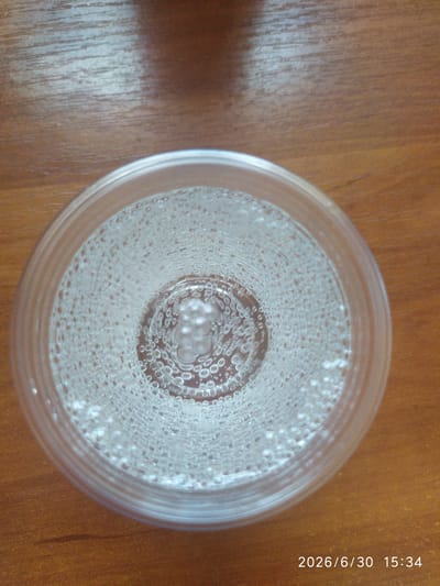 A close-up, overhead shot shows a clear plastic cup filled with a bubbly beverage, likely soda or sparkling water, resting on a wooden surface. The drink is effervescent, with numerous small bubbles clinging to the sides of the cup and rising to the surface. The wooden surface appears to be a table with a visible grain pattern. The lighting suggests an indoor setting, possibly during the day, as there is a distinct shadow cast by the cup to the left. The overall scene is simple and focuses on the texture and visual effect of the carbonated drink.
Visible text in the image reads "2026/6/30 15:34". This indicates the date and time the image was captured, implying it was taken on June 30, 2026, at 3:34 PM. The location context of Skala-Podilska, Ukraine, is provided but not directly evident from visual cues within the image itself.