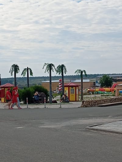 The image depicts an outdoor scene with several artificial palm trees, suggesting a theme park or recreational area, possibly with a beach or tropical theme. In the foreground, three individuals wearing red tops and red headwear are walking from left to right. One person appears to be wearing shorts, and another is wearing pants. Behind them, a small red and yellow building with a peaked roof is visible. To the right of this building, a striped lighthouse structure stands. Further in the background, a large inflatable water slide complex in red and yellow is partially visible, along with other structures that appear to be buildings or stalls. The ground is a mix of paved areas and sandy or gravelly surfaces. The sky is overcast with a pattern of scattered clouds. The location is identified as Sol-Iletsk, Russia.