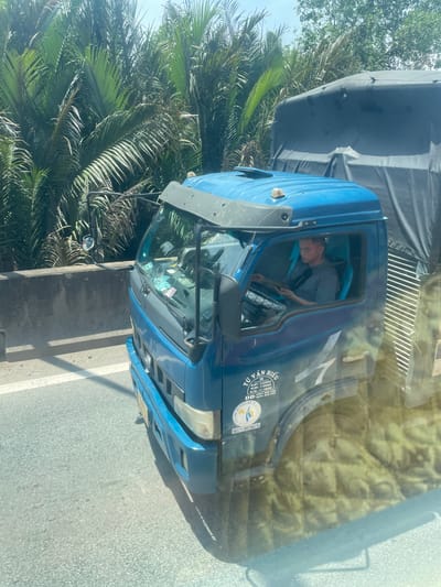 A blue truck is driving on a road in Dong Nai City, Vietnam. The truck has a black tarp covering its cargo. A man is driving the truck and looking at a tablet. The road is lined with palm trees and a concrete barrier. The sky is clear and sunny.
