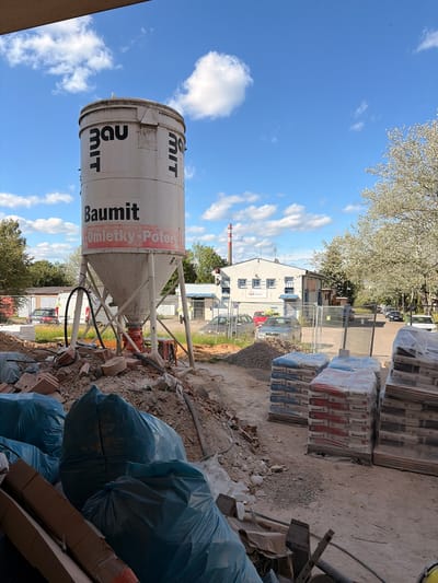 This is a full-frame, eye-level, outdoor, daytime, slightly overcast, full shot of a construction site in Příbram, Czechia. In the foreground, to the left, are several large blue plastic bags of debris. To the right, a large white silo with "Baumit" and "Omítky-Potěry" written on it stands on a metal frame. Below the silo, a pile of construction debris including bricks and rubble is visible. Stacked to the right of the silo are pallets of bagged construction materials, some wrapped in plastic. In the background, there are trees, a building with a red and white striped smokestack, and several parked cars. The sky is blue with scattered white clouds.