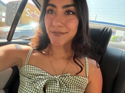 A medium shot captures a woman with dark hair and brown eyes wearing a green and white gingham top. She is in the driver's seat of a car. Outside the car window, a sign for "Sabritas" is visible, indicating the presence of this brand in the local environment, which appears to be in Santa Ana Xalmimilulco, Mexico.