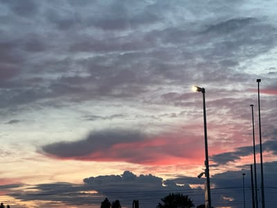 This image depicts a cloudy sky with hues of pink and gray, indicative of a sunset or sunrise. Several lampposts and wires are visible in the foreground, silhouetted against the sky. One lamppost is illuminated, casting a warm glow. Trees can be seen at the bottom of the image, suggesting an outdoor setting, likely in an urban or suburban area. The presence of lampposts and wires hints at infrastructure typical of a developed area. The sky's coloration and cloud formations suggest a specific weather phenomenon. The scene appears to be captured from a ground-level perspective, looking upwards. Without further context, a precise location within Laagri alevik, Estonia, cannot be determined, but the elements present are consistent with such a location.
