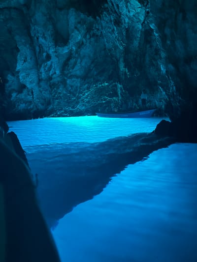 The image captures the interior of a cave, characterized by a dominant blue luminescence. The cave walls are rough and textured, with visible striations and formations. The water within the cave is a vibrant, glowing blue, reflecting the light from an unseen source, likely sunlight filtering through an opening or an artificial light source. A small blue boat is present in the water, with the number "240-KZ" and text reading "NAUTIČKI CENTAR KOMIŽA" visible on its side. A person stands in the boat, facing away from the camera. The overall scene suggests a natural phenomenon of light interaction with water and rock, typical of sea caves. This location is likely the Blue Cave (Modra špilja) near the town of Komiža on the island of Vis, Croatia, a well-known tourist attraction.