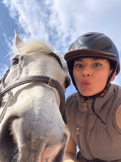 The image is a low-angle selfie featuring a woman wearing a black equestrian helmet and a gray padded vest, making a kissing face. Below her is a gray horse with a black bridle. The background reveals a blue sky with light clouds. The location, Grad Labin, Croatia, cannot be determined from this image.