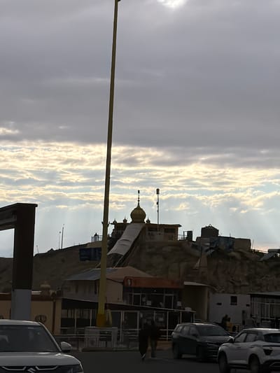 The image depicts a view of the Chumkar Monastery in Chumkar, India, under a cloudy sky. A tall yellow pole stands prominently in the foreground, bisecting the frame vertically. The monastery, situated on a hill, features golden domes and various structures with weathered appearances. Several cars are parked on a street in front of the monastery, and two individuals are walking on the sidewalk. The overall scene is characterized by a slightly overcast sky with visible light rays penetrating through the clouds.