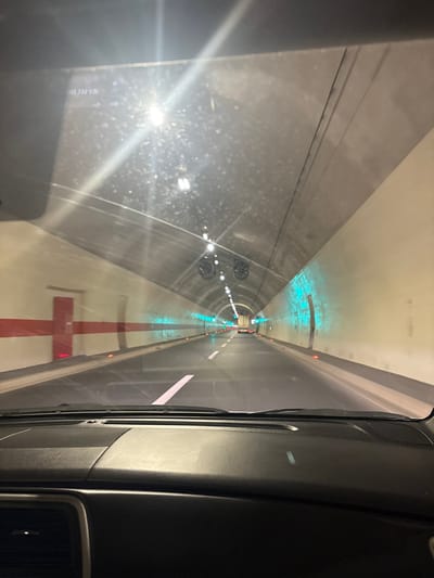 The image captures a forward-facing view from inside a vehicle traversing a two-lane road tunnel in Donje Mrke, Montenegro. The tunnel features an arched, light-colored concrete ceiling with overhead white lighting and multiple circular ventilation fans. The walls are predominantly light-colored. The left tunnel wall presents a red horizontal stripe intersecting with a vertical red stripe, forming a cross pattern, adjacent to a red emergency door. The right tunnel wall contains segments illuminated by blue-green lights and an observable alcove further down the tunnel. The roadway is dark asphalt with white solid and dashed lane markings. In the far distance, a white truck with illuminated red taillights is visible. The lower portion of the image shows the vehicle's black dashboard. Reflections of the tunnel lights are present on the windshield, which also bears a faint digital overlay "H3.702.026" in the upper left corner.