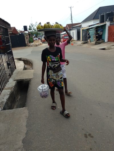 The image shows a street scene in Ohokobe Ndume, Nigeria. A young person walks down the street carrying a tray of bananas on their head and a clear plastic container in their hand. They are wearing a patterned dress and sandals. Behind them, another young person in a pink shirt carries something on their head. The street is paved and lined with buildings, including houses with gates and a small storefront.