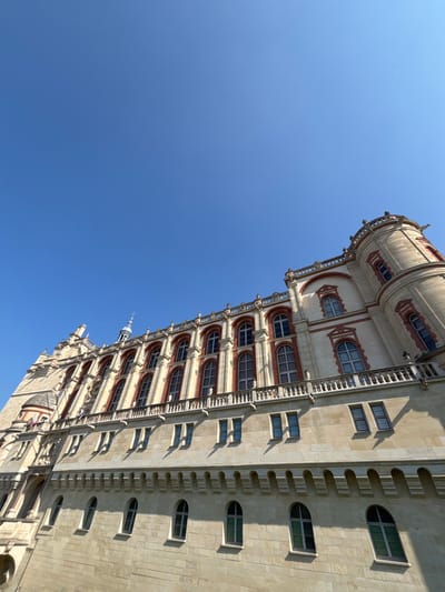 The image displays a detailed view of the Château de Saint-Germain-en-Laye, France, under a clear, bright blue sky. The stone structure, rendered in light-colored masonry with reddish-brown brick accents, rises prominently from the bottom of the frame, viewed from a low angle. Multiple architectural levels are visible: a lower section features robust, light-colored stone with small, arched windows. Above this, a story with smaller rectangular windows casts distinct shadows on the facade due to strong sunlight. The upper levels are characterized by tall, arched windows framed by reddish-brown brickwork and topped with white decorative elements, situated beneath a balustraded balcony running horizontally across the building. A round tower, or turret, with similar arched windows and an ornate top, is positioned on the far right. A smaller, pointed spire is visible along the central roofline. No individuals or activities are present within the frame.