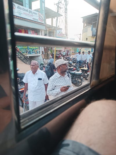An outdoor scene in Kothacheruvu, India, is captured from the interior of a vehicle, framed by a dark window frame. The image depicts a bustling street with several individuals, parked motorcycles, and multi-story commercial buildings under bright daylight.
In the immediate foreground, outside the vehicle's window, an elderly man with short grey hair and a white mustache stands facing left. He wears a short-sleeved white shirt with a smartphone visible in his left breast pocket, and white trousers. His right hand holds a light-colored walking stick, and his left hand carries two blue shopping bags. To his right, a man with a beard and glasses wears a light-colored plaid shirt and a beige cap, looking right while holding a white smartphone in his right hand.
The background features numerous parked motorcycles and scooters lining the street. Several men are visible further back; one group of three men stands near the motorcycles, with one appearing to wear a light blue uniform. Behind the elderly man, a person with a dark head covering and a black backpack is visible.
Buildings line the street. On the left, a multi-story structure displays a prominent sign in Telugu script for "తేజ దంత వైద్యశాల Dr.P.V.SreeTeja BDS (Dental Surgeon)" on its upper floor. Below this sign, various garments are hung for display. Further down the street, additional commercial signs in Telugu are visible. To the far left in the distance, a tall utility tower rises. The street surface appears to be paved, with some dirt visible. The bottom right of the image is partially obscured by the blurred leg of the person inside the vehicle from which the photograph was taken.