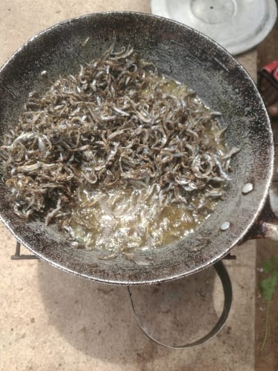 The image shows a high-angle view of a dark, worn metal pan, possibly a wok or large skillet, filled with numerous small fish being fried in oil. The pan appears to be old and well-used, with a dark, textured, and somewhat mottled surface. Several silver rivets are visible on the inner side of the pan near the rim.

The small fish, which are slender and curled, are a mix of silver, grey, and dark brown hues. They are densely packed in the pan, forming a substantial pile. A significant amount of cooking oil is visible at the bottom of the pan, actively bubbling and glistening, indicating that the fish are being cooked over heat. The oil appears a light golden-yellow color, and some of the fish are submerged in it, while others are piled higher, resting above the oil level.

The pan is placed on a simple, dark metal stand or ring, which is partially visible supporting the pan from underneath. This suggests outdoor cooking or a rustic kitchen setup. The surface beneath the pan and stand is a rough, light-colored material, likely concrete or packed earth, with some shadows cast upon it.

In the upper right background, a blurred, light-colored metal lid, possibly from another pot, is partially visible. Further to the right, out of focus, there's a small hint of red and a blurry green patch, possibly vegetation. The lighting suggests it is daytime. No text is visible in the image.