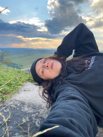 A young woman with dark hair and a black beanie lies on a rock overlooking a vast, green landscape. She is wearing a black hoodie with white lettering on the sleeve. The sky above is filled with dramatic clouds, with rays of sunlight breaking through. The foreground shows the rocky terrain and some green plants. The location is in the village of Абзаново, Russia.