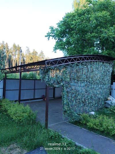 A carport with a metal frame and a corrugated metal roof is covered with camouflage netting on one side. The carport is situated in a yard with lush green grass and bushes. A tall fence made of corrugated metal is visible behind the carport. In the background, there is a dense forest with tall pine trees and a clear blue sky above. The image also shows a paved walkway in front of the carport and a portion of a residential building. The bottom right corner of the image displays text which appears to be a timestamp: "realme 12 21 июня 2026 г. в 20:17".