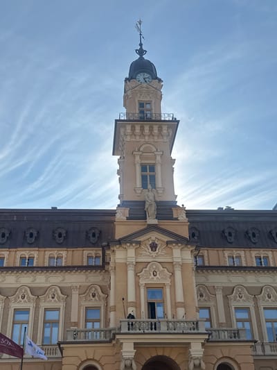 The image shows the facade of the historic Town Hall in Nowy Sącz, Poland. The building is constructed in a light-colored stone and features ornate architectural details, including columns, sculpted figures, and decorative window surrounds. A prominent clock tower rises from the center of the building, topped with a weather vane. The sky above is a bright blue with wispy white clouds. In the foreground, a balcony is visible with a single person standing on it, looking out. Flags are also present on the left side of the building, and some architectural elements suggest it is daytime. The overall impression is one of historical significance and architectural grandeur.