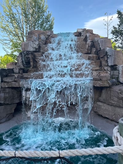 This media file presents a vibrant man-made waterfall feature, located in Chelsea, Canada, during daylight hours. The central subject is the waterfall itself, where turquoise water cascades over dark, angular rock formations, appearing to be columnar basalt. The water splashes vigorously at the base, creating abundant white foam and a dynamic, frothy appearance.

The scene is framed by natural elements. On the left and top, lush green foliage from both coniferous and deciduous trees is visible against a bright, clear blue sky, which features a few wispy white clouds. The rocks are predominantly dark grey and brown, contributing to a rugged, naturalistic aesthetic.

In the foreground on the right side of the image, a thick, white rope with a visible texture runs diagonally downwards, suggesting it is part of a barrier, railing, or nautical-themed structure, possibly alongside a viewing platform or pathway. A dark net or mesh is partially visible behind the rope.

The time of day is clearly daytime, indicated by the bright, even lighting and the clear sky. The weather appears sunny and pleasant. The overall atmosphere evokes natural beauty and the refreshing power of moving water. No people or visible text are present in the image.
