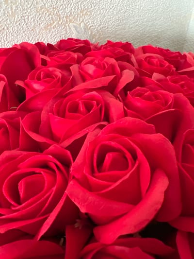 The image displays a close-up view of a bouquet of vibrant red roses. The roses are arranged densely, with many petals overlapping and obscuring the centers of some flowers. The background is a textured, pale surface, possibly a wall or fabric, which is out of focus. There is no indication of the location being Heorhiivka, Ukraine, nor are there any people, actions, or interactions depicted. The focus is solely on the texture and color of the rose petals.