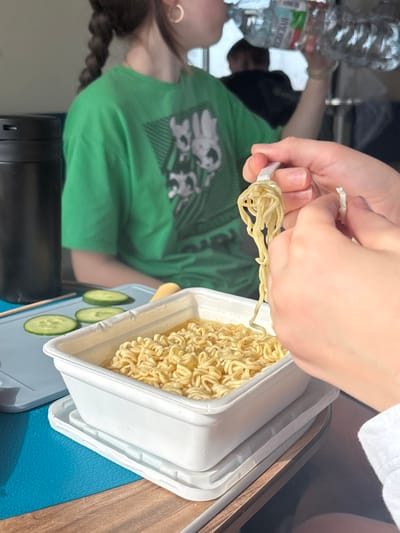 The scene depicts an indoor setting, likely a dining area, in Karmanovo, Russia. In the immediate foreground, a pair of hands is active; the left hand holds a white plastic fork, from which a portion of yellow instant noodles is lifted, while the right hand holds a small, irregular piece of white plastic, possibly packaging. A white rectangular container of instant noodles rests on a white plastic tray, situated on a blue mat. To the left of the noodles, three circular slices of green cucumber are arranged on a lighter blue surface. A tall, black cylindrical thermos or bottle stands upright to the far left. In the midground, partially obscured, a person with dark hair styled in braids, wearing a green t-shirt with a dark graphic print, is drinking from a clear plastic bottle with a white cap, which displays Cyrillic text on its label. A gold hoop earring is visible on this person's left ear. Another indistinct figure is partially visible in the blurred background. The setting includes a light brown wooden table surface.