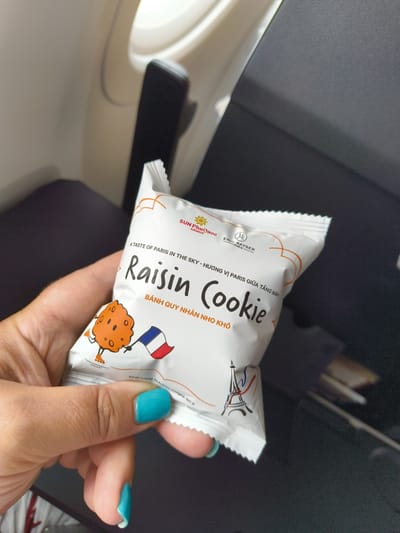 A person's hand holds a small, individually wrapped cookie in front of a dark gray airplane seat. The cookie packaging is white with orange and black text and graphics. The text includes "SUN PHU QUOC AIRWAYS," "A TASTE OF PARIS IN THE SKY," "Raisin Cookie," and Vietnamese text. There is an illustration of a cartoon cookie with roller skates and a French flag on the left side of the packaging, and an illustration of the Eiffel Tower on the bottom right. The person's fingernails are painted a bright teal color. The background appears to be the interior of an airplane cabin, with a window on the left side and the back of a seat on the right. The scene is indoors and dimly lit, suggesting it is inside an airplane.