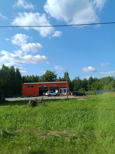 The image shows an orange building with a dark roof and several windows, including a large round window. Two cars are parked in front of the building. One is a white SUV, and the other is a dark sedan. There are a few people standing near the white SUV. The building is surrounded by trees and a grassy field. The sky is blue with scattered white clouds, and a power line is visible in the upper part of the image. The scene appears to be in a rural or suburban area, possibly a commercial establishment like a car wash or repair shop.