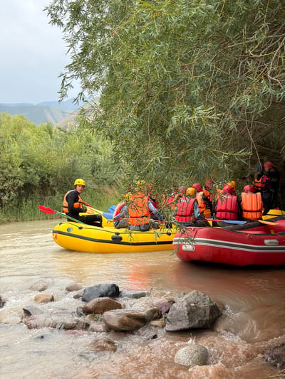 This media file depicts a group of people in a river rafting activity in Sasyk-Bulak, Kyrgyzstan. Two inflatable rafts, one red and one yellow, are partially submerged in the muddy water. The rafts are filled with passengers wearing life vests and helmets in various colors, including orange and red, suggesting a recreational or guided tour. One individual in the yellow raft is wearing a yellow helmet and an orange life vest, holding a red paddle and looking towards the camera. Trees with green foliage line the bank of the river, and in the background, there are hints of hills and possibly a cloudy sky, indicating it might be daytime. The water appears somewhat turbulent, with visible rocks along the riverbed. The overall impression is of an active outdoor adventure in a natural setting.
