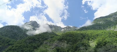 A wide shot captures a lush green mountain range under a bright blue sky dotted with white clouds. The mountains are heavily forested with dense green trees, and rocky outcrops are visible in places. Wisps of white fog cling to the upper slopes, partially obscuring the peaks and adding a dramatic effect. A thin dark line, likely a cable, is visible in the upper left corner of the frame, suggesting the presence of a cable car or similar structure. The scene appears to be a natural landscape with no visible people or man-made structures other than the potential cable. The image is taken from a low angle, looking up at the imposing mountains.