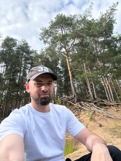 The media file is a selfie taken by a man wearing a white t-shirt and a gray baseball cap with the number "32" in white lettering. He has a beard and is looking at the camera with a neutral expression. The background features a forest with tall pine trees and exposed roots on sandy ground. The sky is visible through the trees, with some clouds present. The lighting suggests it is daytime, possibly late afternoon or early evening, given the angle of the shadows. The location is identified as Severnaya Samarka, Russia, implying an outdoor, natural setting. The man appears to be in a relaxed or casual mood, enjoying the outdoors.