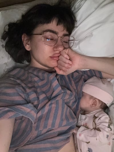 An overhead shot shows a person wearing glasses lying on a pillow with their fist near their mouth. A baby wearing a hat is partially visible to the right, lying on the person's chest. The person is covered with a striped blanket. Both the person and the baby are sleeping. The scene suggests a moment of rest and quiet companionship.
