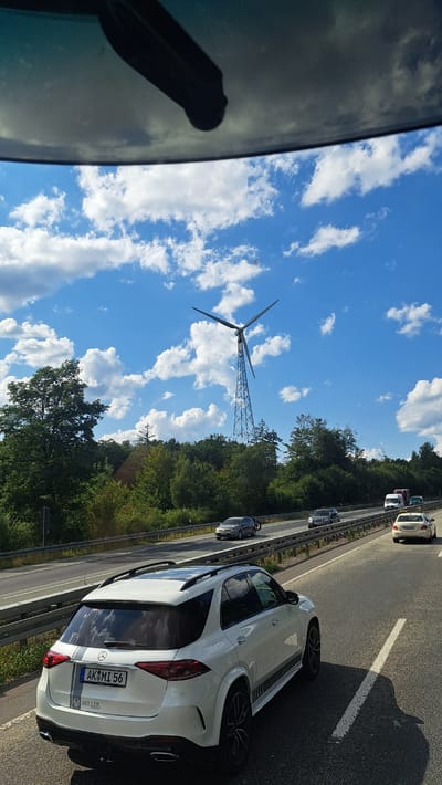 The image was taken on a sunny day, likely from inside a vehicle, looking out onto a highway. The foreground is dominated by the rear of a white SUV. The SUV has a license plate that reads "AK MI 56" and has a stripe design on its side. Other cars are visible on the highway in the middle ground, including a yellow taxi. In the background, a tall wind turbine stands against a bright blue sky filled with white, puffy clouds. The turbine is surrounded by a dense line of green trees. The location is the city of Freudenberg, Germany.