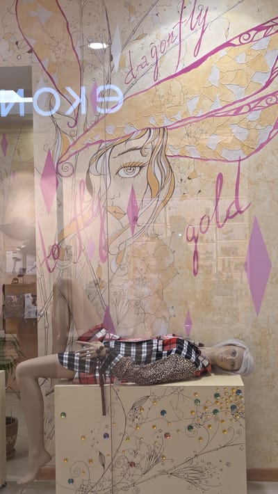 The image shows a mannequin lying on top of a decorated cabinet in what appears to be a shop window display. The mannequin has light-colored hair and is wearing a black and white plaid dress with leopard print accents. The cabinet is light brown and adorned with swirling line art and colorful gem-like embellishments.

Behind the mannequin, a large illustration is visible on the glass. This illustration features a stylized drawing of a woman's face, with large eyes and flowing hair. Above the woman's head, large wings resembling those of a dragonfly are depicted, with the text "dragon fly" written in a script font. The word "gold" is also visible in a similar script, positioned to the right of the illustration.

On the left side of the glass, the reflective surface shows part of the interior of a store, including what appears to be signage with Cyrillic letters, possibly reading "ЕКО" or similar. The background of the illustration has a textured, marbled appearance in shades of beige and pink.

The overall scene suggests a fashion display with a fantasy or whimsical theme. The location is identified as Novotlitsy, Russia, implying this is a storefront in that city.