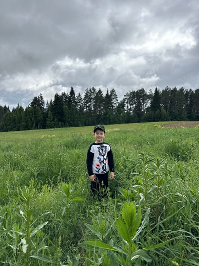 An image captured in Романята, Russia, depicts a young male child standing centrally in a dense, tall grassy field under an overcast sky. The child, facing the viewer, wears a black and white long-sleeved shirt featuring a stylized lion's head and floral patterns, dark trousers, and a dark baseball cap with a subtle emblem. His facial expression is a slight smile. The field is dominated by various green vegetation, including tall grasses and broad-leafed plants, extending from the foreground to the mid-ground. In the background, a dense line of coniferous and deciduous trees forms a dark green barrier, stretching across the horizon. The sky is completely covered by thick, grey, stratiform clouds, with some lighter areas indicating variations in cloud density.