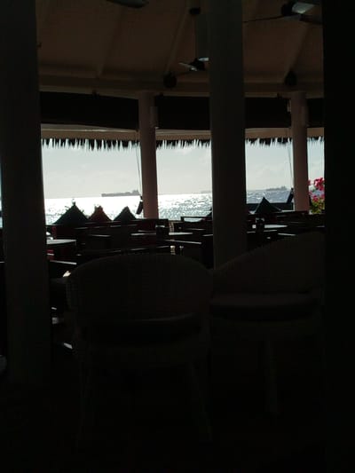 An outdoor seating area with thatched roofs and dark furniture is positioned in front of a body of water. The sun is shining brightly, creating a shimmering reflection on the water's surface. Several large ships are visible in the distance on the horizon. The scene appears to be located in Gulhi Falhu, Maldives, a developing industrial island in the South Malé Atoll.