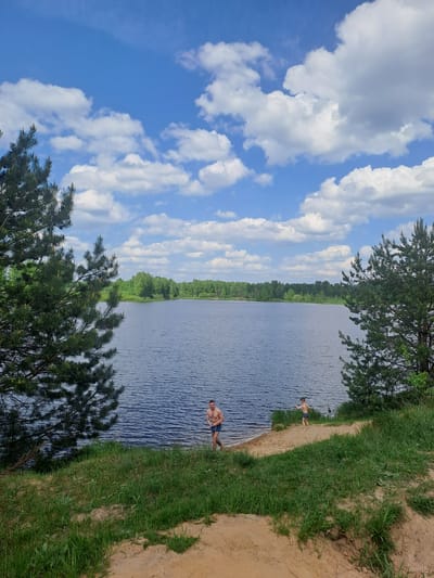 The image depicts a body of water, possibly a lake or river, with two individuals visible on the shore. One person, appearing to be an adult male, is standing in the water near the edge. Another person, possibly a child, is further along the shoreline. The background shows a lush green forest lining the opposite bank. The foreground features a sandy bank with grass, and trees frame the scene. The sky is blue with numerous white clouds. Given the prompt, the location is Avdot’ino, Russia.